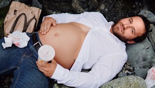 A new dad in a white half-buttoned shirt with his belly sticking out and food next to him, doing a "...
