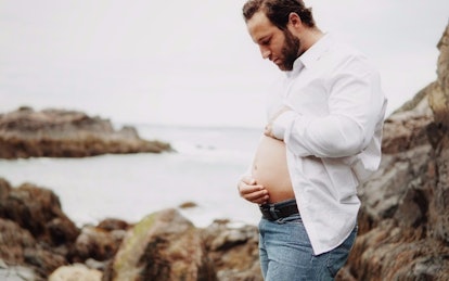 A new dad in a white half-buttoned shirt, holding his belly on a rocky beach by the sea, doing a "ma...
