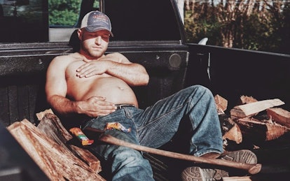 A topless new dad, holding his belly with an axe and chopped wood next to him, doing a "maternity" p...