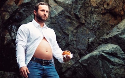 A new dad in a white half-buttoned shirt with his belly sticking out holding a small burger in his h...