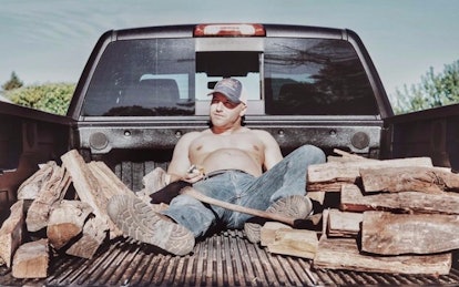 A topless new dad, holding his belly, lying in the trunk of a pickup truck with an axe and chopped w...