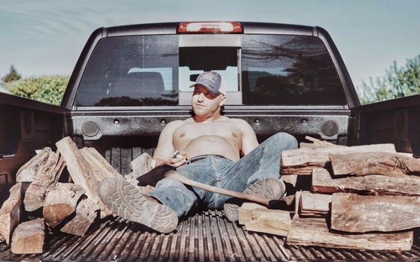 A topless new dad, holding his belly, lying in the trunk of a pickup truck with an axe and chopped w...