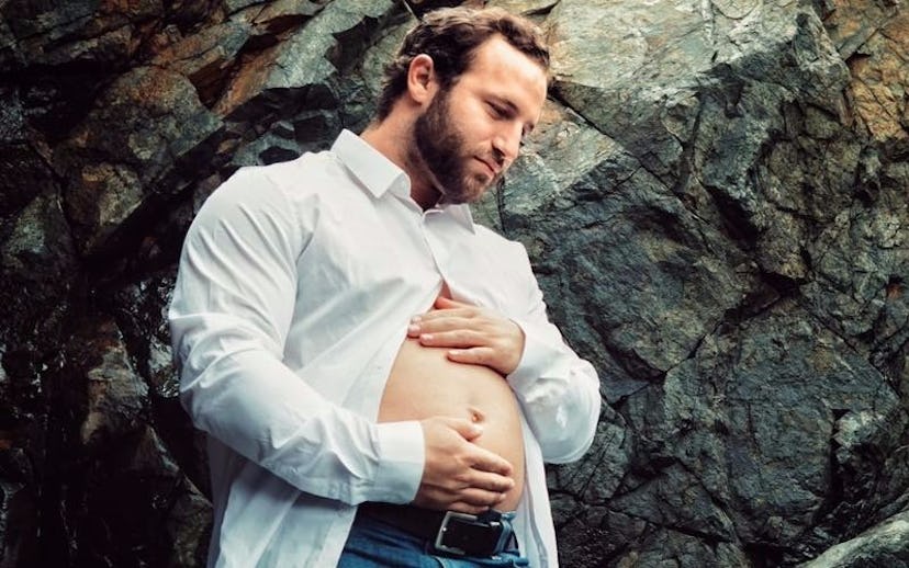 A new dad wearing a white half-buttoned shirt holding his belly with a rocky background, doing a "ma...
