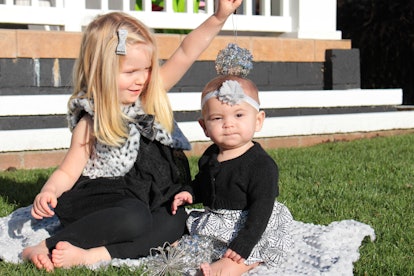Two baby girls sitting outside on grass and smiling while dressed up with bows on their heads