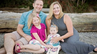 Mother and father with two kids smiling and sitting on the ground, holding a photo of a baby they've...