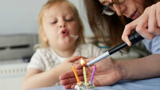 Mother lighting candles on a cupcake and covering it with her other hand, and a child blowing air be...