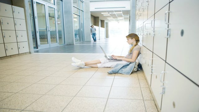 A girl sitting on the floor in front of lockers in a school while doing her homework on her laptop