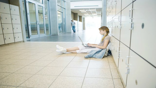 A girl sitting on the floor in front of lockers in a school while doing her homework on her laptop 