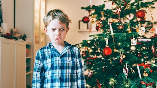 Sad and angry blonde boy wearing a blue and white pajamas standing next to a Christmas tree with red...