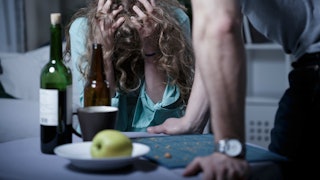 A woman holding both of her hands on her head next to a man who is approaching her at a dining table