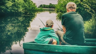 Grandfather and grandchild fishing in a green boat on a lake surrounded by water and trees