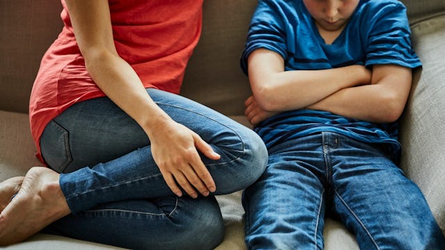 A mother wearing a red shirt and blue jeans talking to her angry son with crossed arms while sitting...