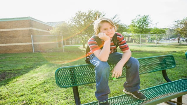 A boy sitting on the top of a bench placing one hand on his cheek.