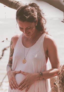 A smiling pregnant dark-haired curly woman with her hair tied up in a white tank top holding her han...