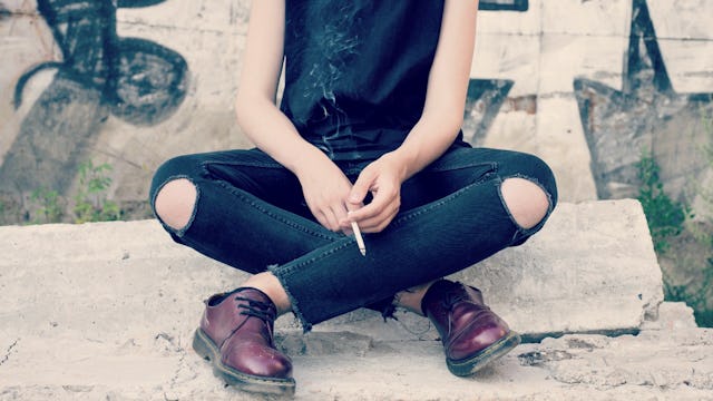 A teen dealing with addiction sitting cross-legged holding a cigarette in a black t-shirt, ripped je...