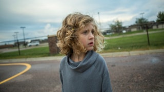 A wavy-haired transgender boy in a grey sweater in a basketball field looking left