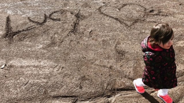 A little girl walking on the beach in a raincoat and pink shoes with "Lucy" and a heart written on t...