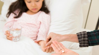 A mother handing ADHD medication to her daughter, who is laying in bed and holding a glass of water.