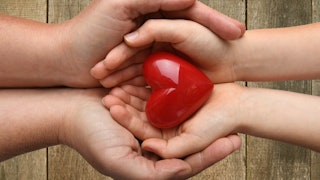 A mother clasping her child's hands, within which rests a red heart.