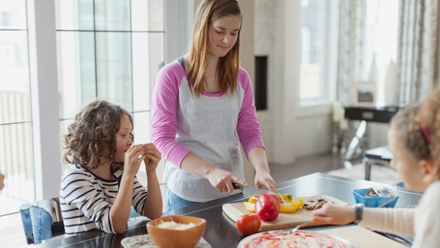 A babysitter chopping veggies on a board next to two girls in the kitchen.