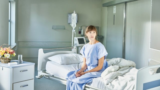A woman with bangs in a light blue hospital gown sitting on the edge of a bed after her miscarriage