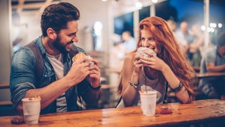 A boy with brown hair and beard and a girl with red hair eating burgers at a restaurant and laughing