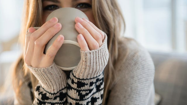 A close-up of a woman drinking from a grey cup while wearing a beige sweater