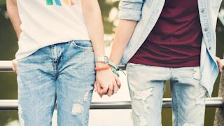 Two guys wearing blue jeans and white and red shirt, holding hands while leaned back on a metal fenc...