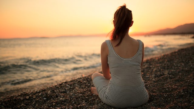 A woman wearing a white and blue striped dress sitting on the beach watching the sunset
