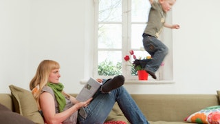 A woman in a grey shirt and green scarf reading a book while her kid is jumping off the couch 