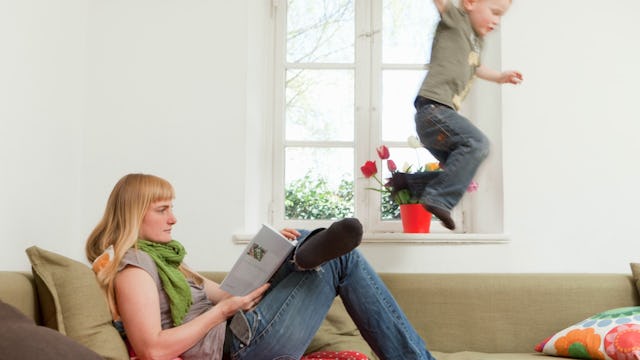 A woman in a grey shirt and green scarf reading a book while her kid is jumping off the couch