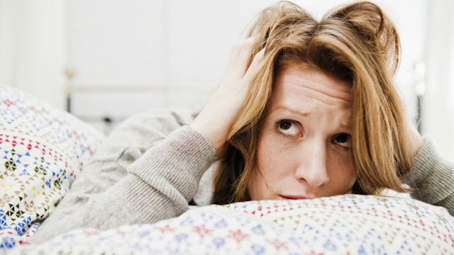 A woman with Generalized Anxiety Disorder in a grey sweater lying in a bed on her stomach, holding h...