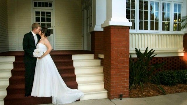 A man in a suit and a woman in a white wedding dress holding a bouquet of flowers while kissing on t...