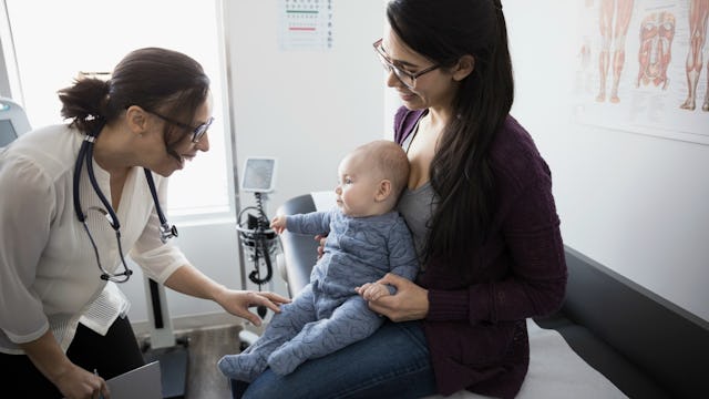 A woman in a black shirt and glasses sitting and holding her baby while a pediatrician is checking t...