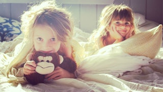 Two little girls laying down together on a messy bed with big smiles on their faces.