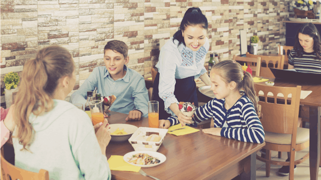 A restaurant server giving an ice cream to a girl who's sitting at a table next to her family