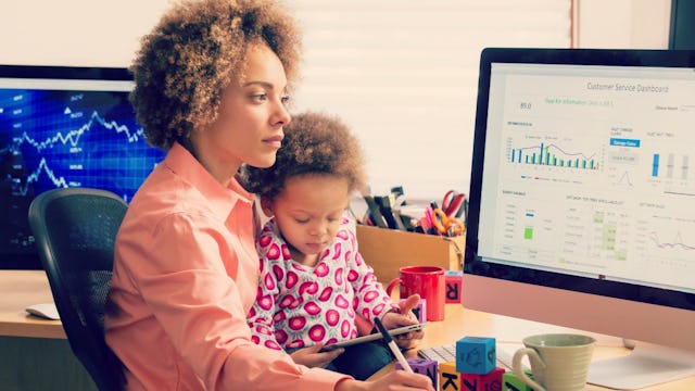 A working mom with curly brown hair holding her child in her lap while simultaneously working on her...