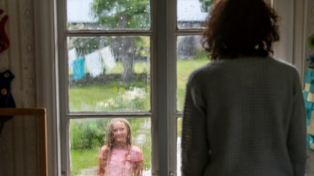 A mother gazes through a window, observing her daughter in the garden, dressed in a pink shirt.