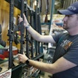 A man wearing a grey cap and grey T-shirt, and a watch picking up a riffle in a gun shop