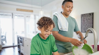 A father in an olive T-shirt and son in a bright green T-shirt washing dishes together and smiling