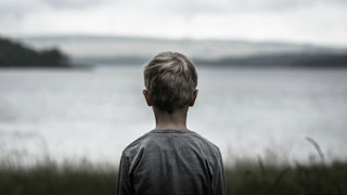 A blonde boy in a grey shirt from the back standing and looking at the sea 