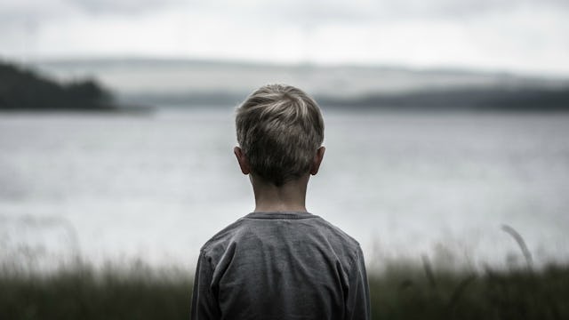 A blonde boy in a grey shirt from the back standing and looking at the sea