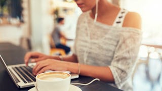 A woman typing on her laptop with earplugs in her ear and a cappuccino in front of her