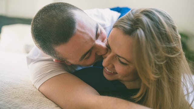 A husband lying down on a bed with his wife, hugging her and kissing her cheeks.