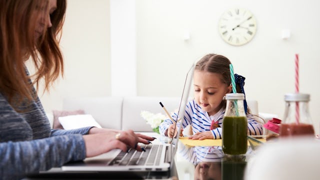 A mom doing her job tasks on a laptop while, at the same table, her daughter is working on her homew...