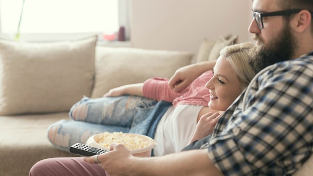 A married couple watching TV and eating popcorn while lying on a couch