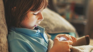 A 5-year-old boy home alone sitting on the couch and playing with his phone