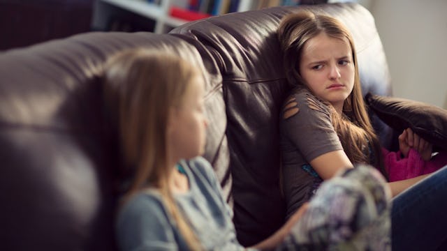 Two angsty tween sisters sitting on a couch and looking at each other with angry facial expressions