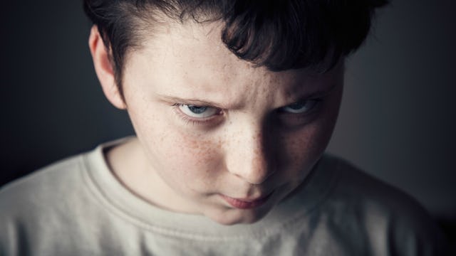 Angry-looking brown-haired boy in a grey sweatshirt looking directly into the camera with a dark bac...