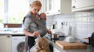 Blonde woman cooking in a kitchen while carrying a baby on her chest and offering a spoon to a littl...